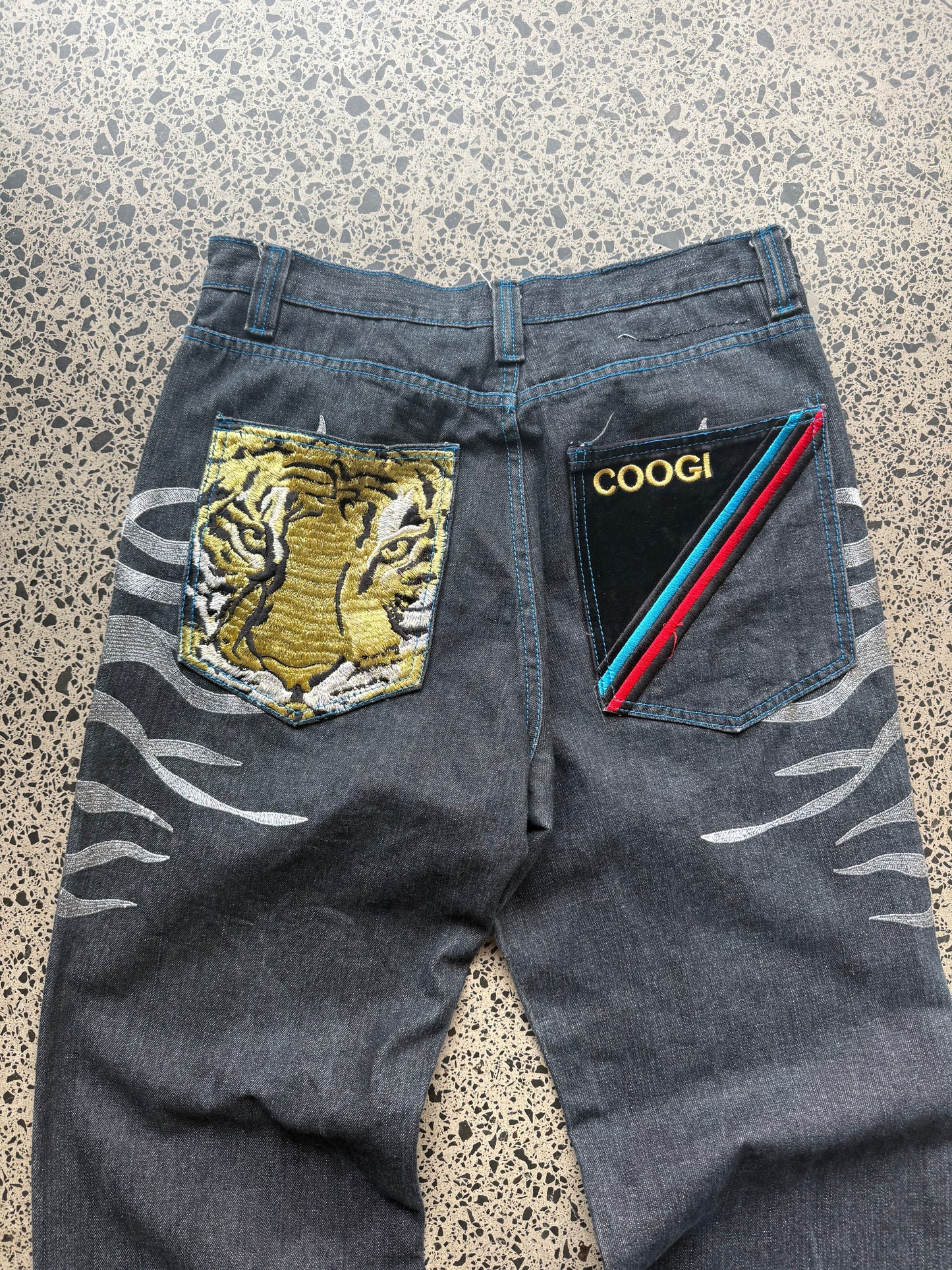 Coogi tiger embroidered denim jeans, 32W x 34L, laid on a polished concrete background — medium-blue denim with bold tiger embroidery and contrast stitching.