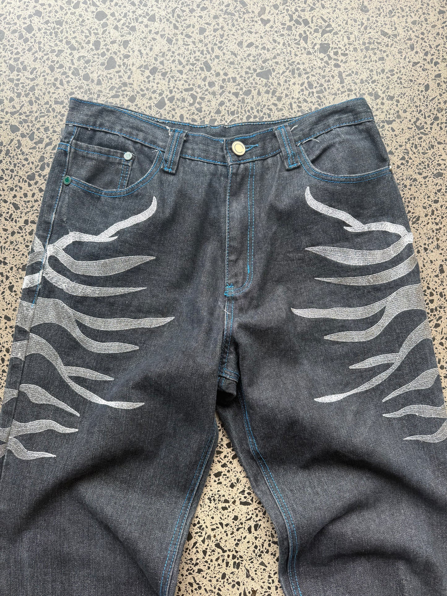 Coogi tiger embroidered denim jeans, 32W x 34L, laid on a polished concrete background — medium-blue denim with bold tiger embroidery and contrast stitching.