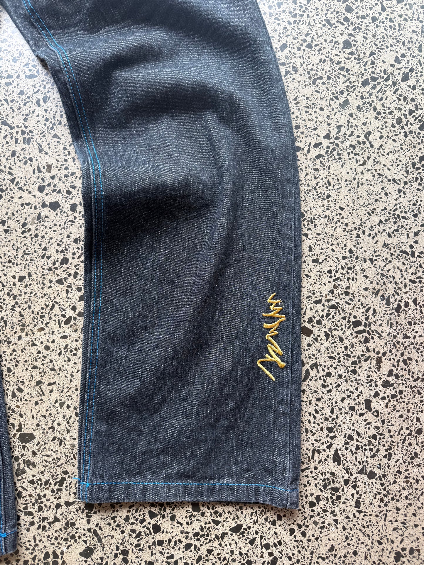 Coogi tiger embroidered denim jeans, 32W x 34L, laid on a polished concrete background — medium-blue denim with bold tiger embroidery and contrast stitching.