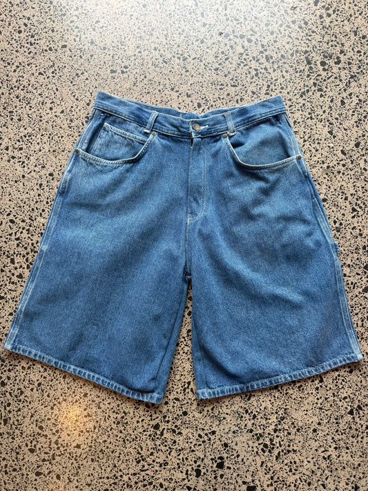 Mid-wash blue wash denim jorts, size 32W, laid on polished concrete background. Knee cut with classic stitching and soft wash