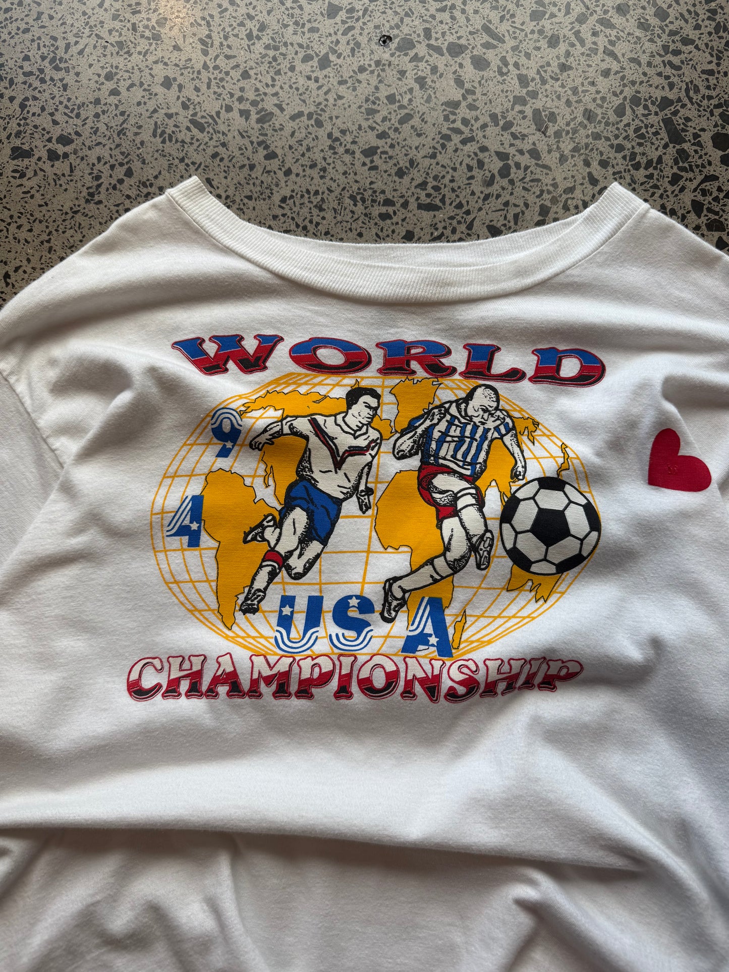 1994 USA World Champs football T-shirt, size L, laid on polished concrete background — vintage red, white, and blue sports graphic on soft cotton.