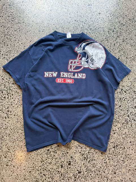 Modern New England Patriots NFL T-shirt in size XL laid on a polished concrete background featuring bold team graphics.