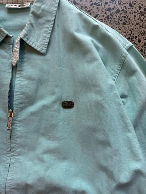 Turquoise Lacoste collared zip-up jacket laid flat on polished concrete background, vintage designer jacket with embroidered crocodile logo and clean fit.