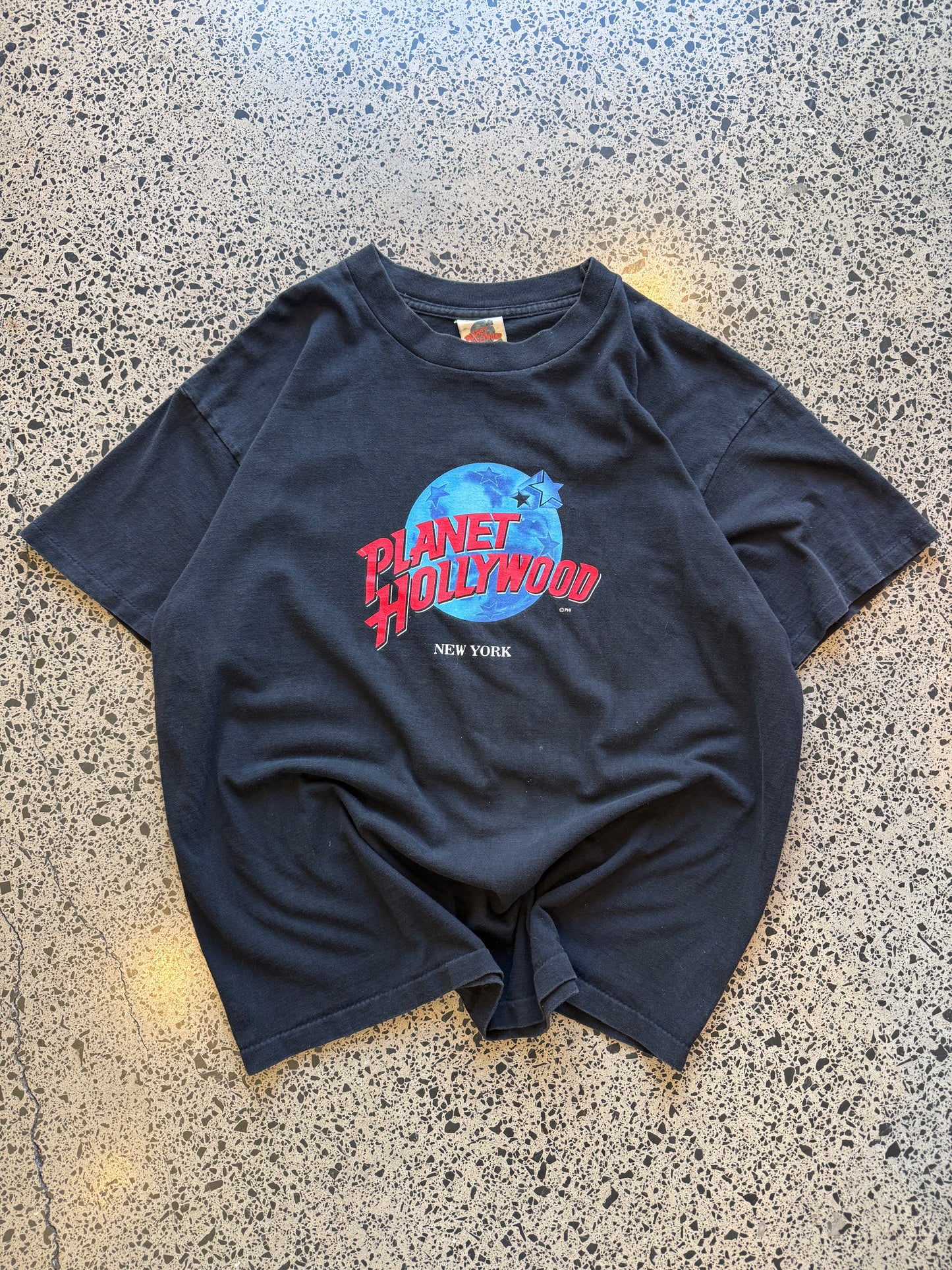 90s Planet Hollywood New York T-shirt in black, size XL, laid on polished concrete background — vintage souvenir tee with bold logo.