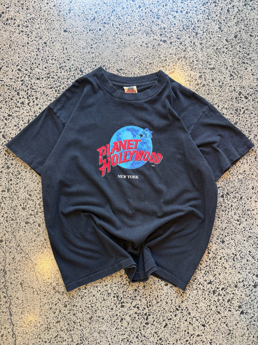 90s Planet Hollywood New York T-shirt in black, size XL, laid on polished concrete background — vintage souvenir tee with bold logo.