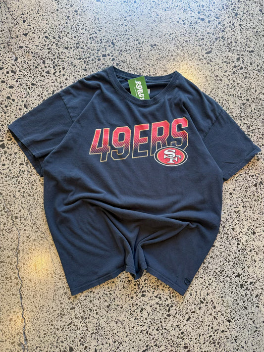 San Francisco 49ers black graphic T-shirt size L laid on polished concrete background — bold red-and-gold team logo on soft cotton.